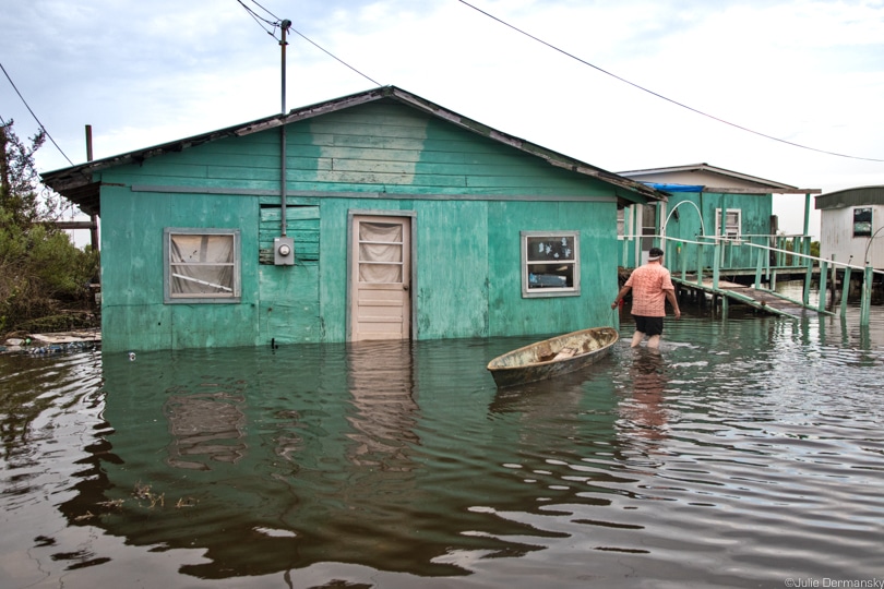 Man in orange t-shirt walks through floodwaters with a small boat, by a bright green boarded-up house
