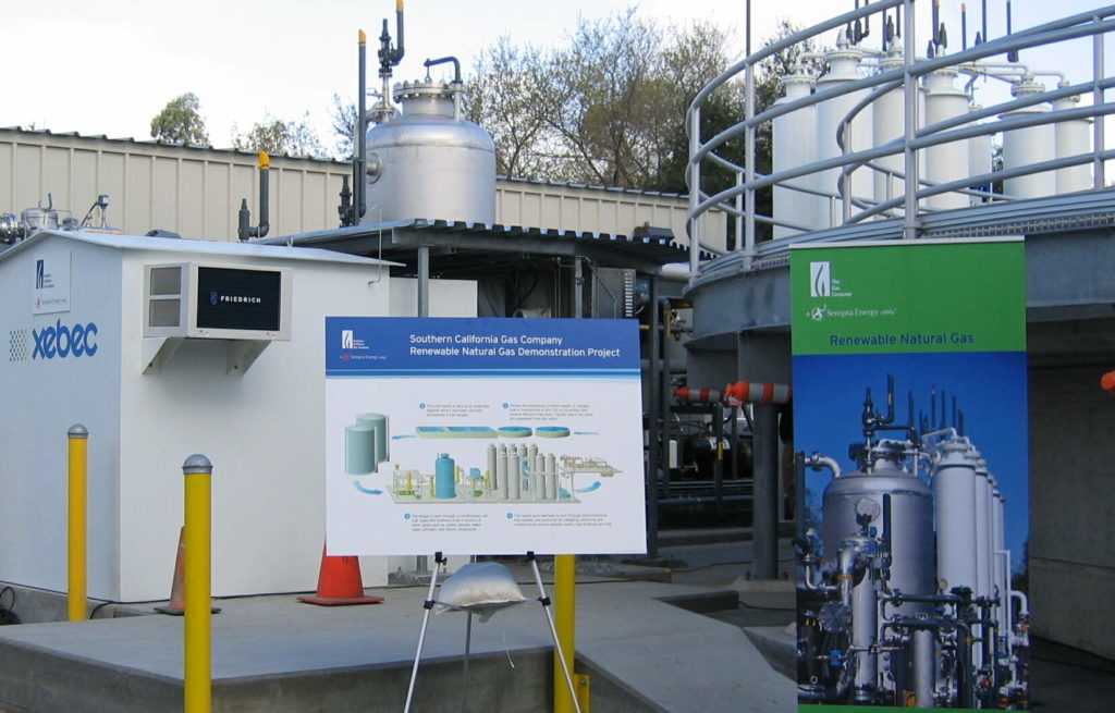 Two signs show information about a SoCalGas renewable natural gas demonstration project, which has a small white building, silver and white tanks in the background.