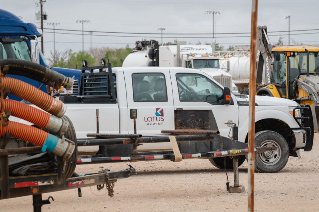 White truck with Lotus logo in an industrial yard with semi trucks, heavy equipment, and vehicles with orange pipes.