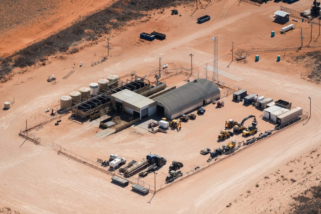 Aerial view of industrial site with rounded metal and white boxy warehouses, large tanks, and heavy equipment in the desert.