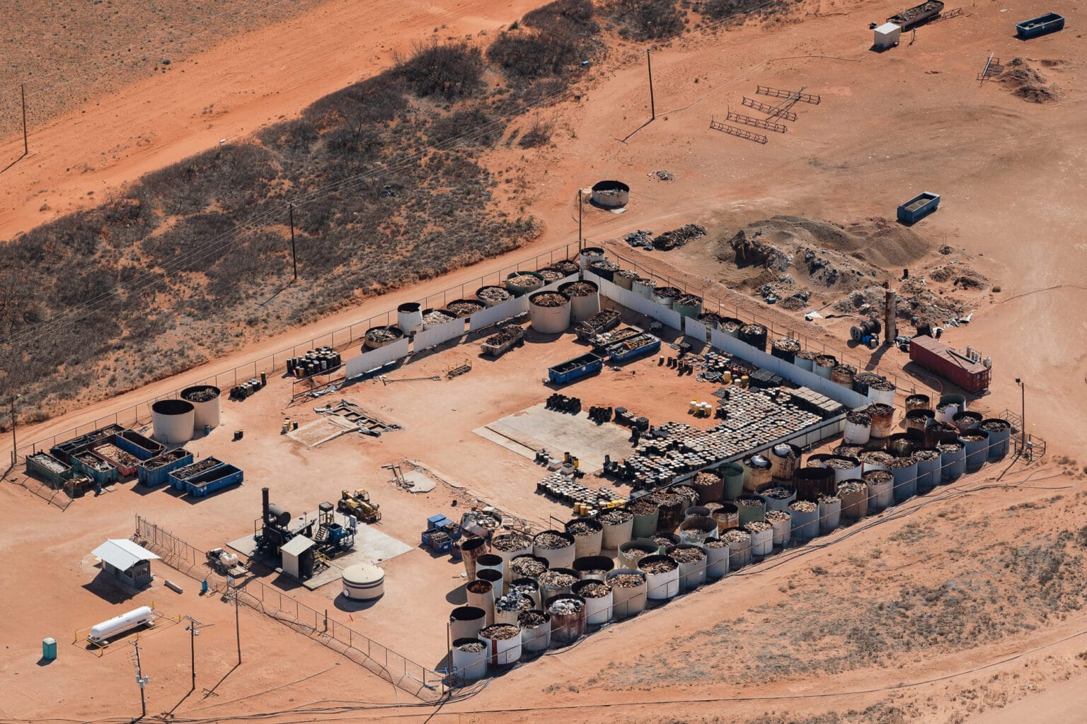 Aerial view of industrial site with dozens of open tanks holding oilfield pipe and other waste and barrels stacked up in the desert.