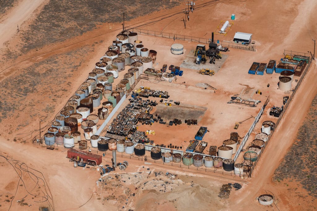 An industrial facility in the desert shows dozens of large tanks filled with pipe and oilfield waste and black barrels.