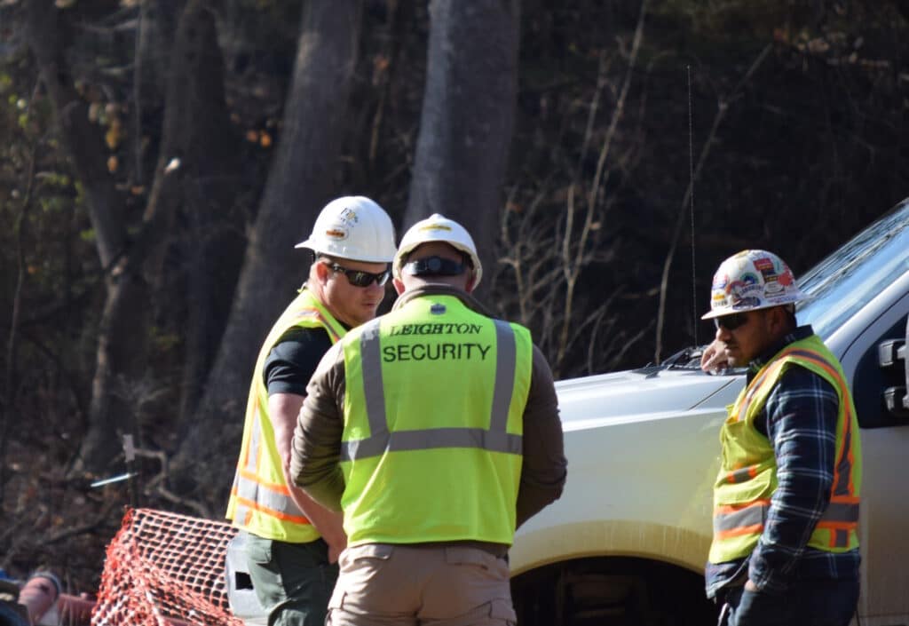 Three men wearing hard hats and yellow security vests that read Leighton Security stand next to a white truck