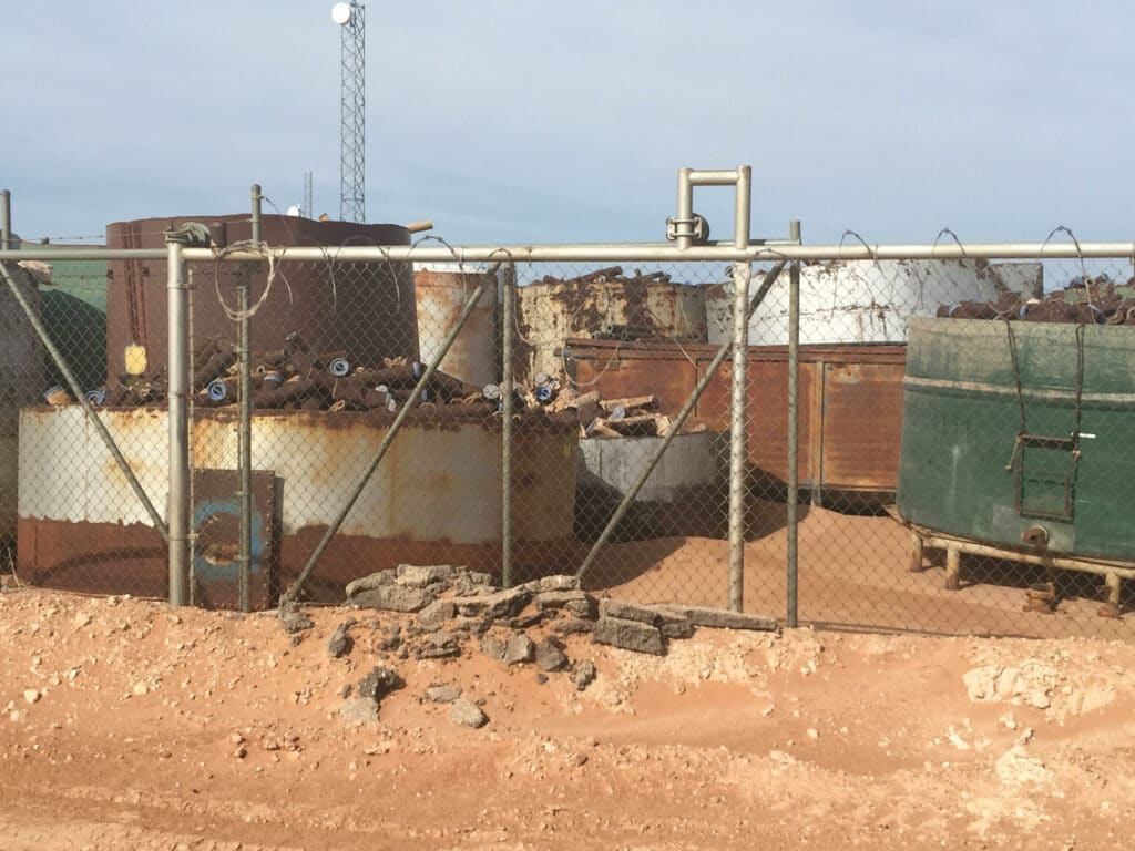 Large rusty, white, and green tanks with rusted pipe sections sit behind a chainlink fence in the red-dirt desert.