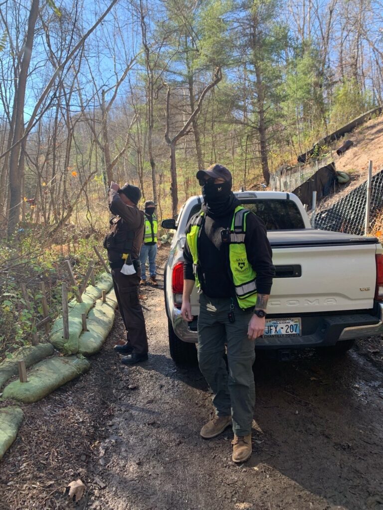 Three men wearing security clothing stand next to a white truck in woods in Virginia