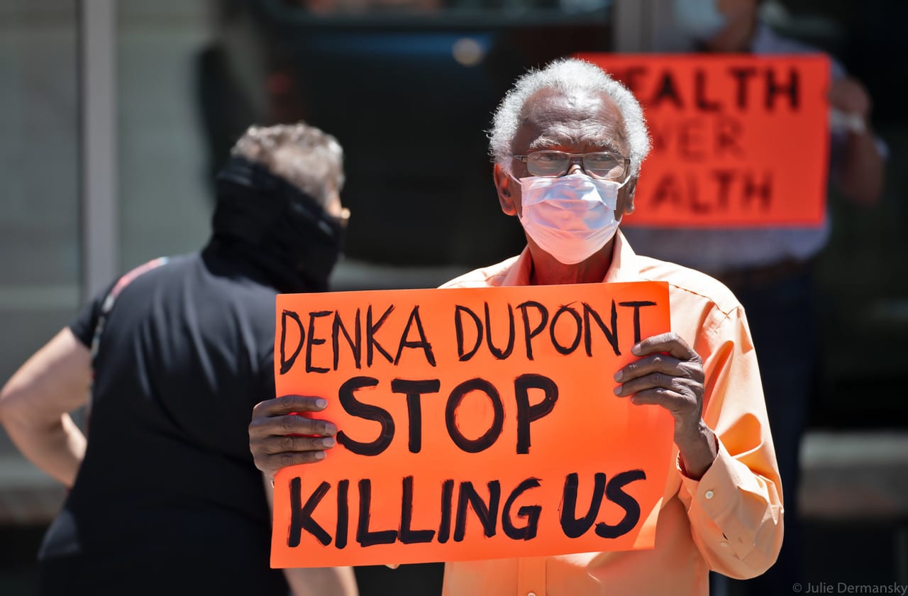An older Black man in a medical mask holds an orange sign with text 'Denka Dupont stop killing us'