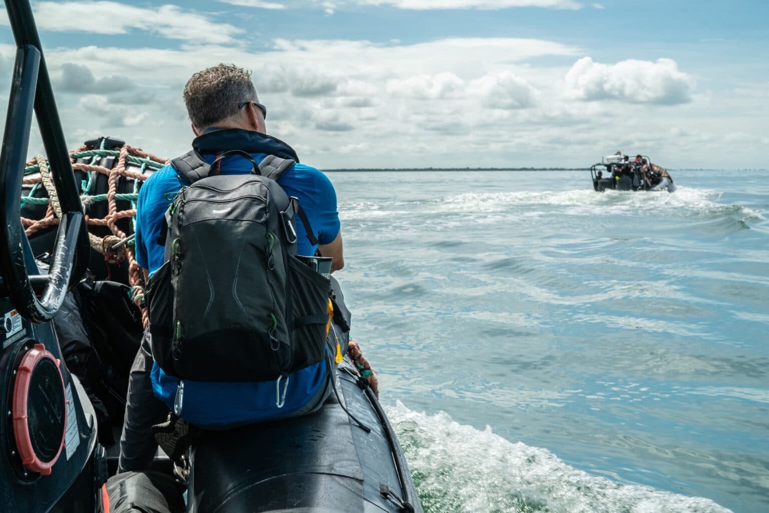 The back of a man wearing a backpack sitting on the edge of a boat and looking across the ocean to another small boat nearby.