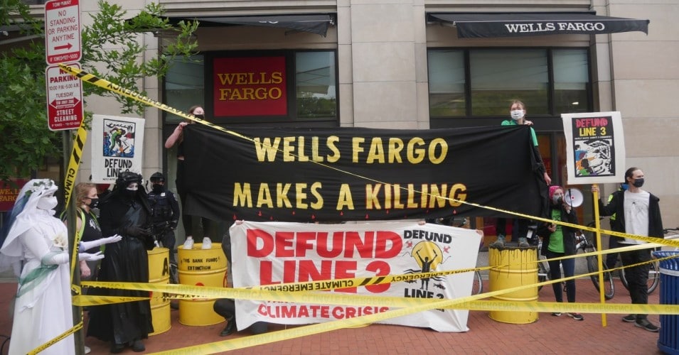 Activists protest banks funding the Line 3 pipeline in front of a Wells Fargo, with yellow caution tape and oil drums and holding banners