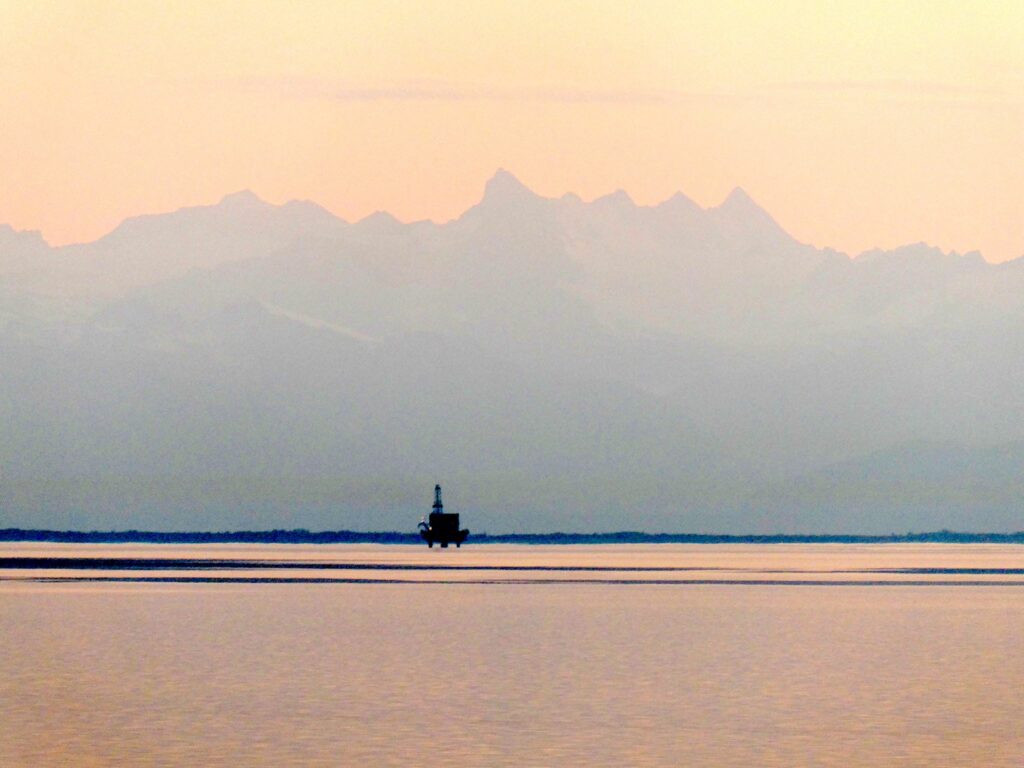 An offshore drilling platform in the distance at sunset with mountains behind.