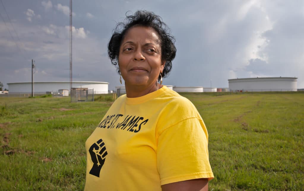 Sharon Lavigne, a Black woman wearing a yellow t-shirt with RISE ST. James and a raised fist, in front of a field of oil tanks