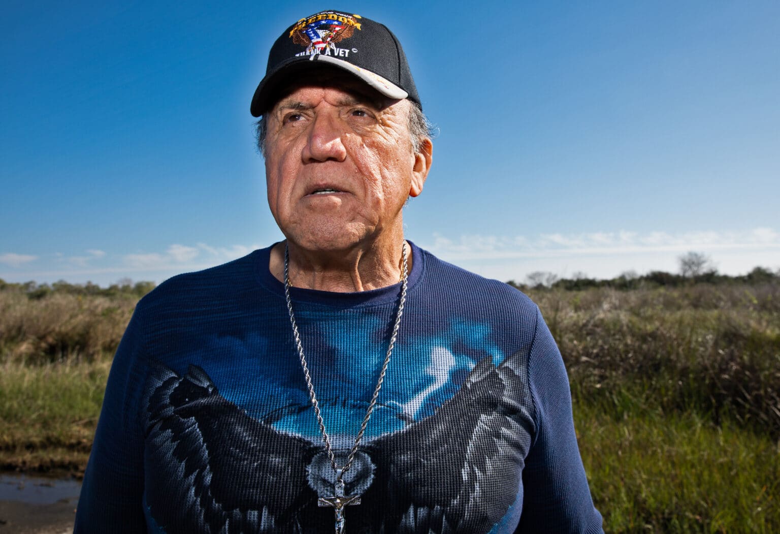 An older man in a baseball cap and blue shirt stands in a marsh and looks away