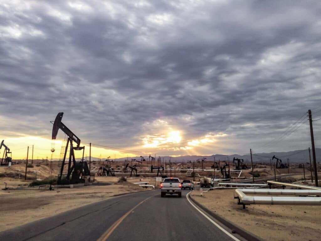 Vehicles drive down a highway through an arid oilfield at sunset