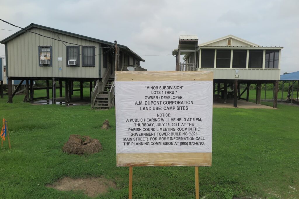 White sign giving notice of A.M. Dupont's proposal to subdivide its parcel of land to enable its sale to fishing camp owners on the Isle de Jean Charles, Louisiana, with two raised houses in the background.