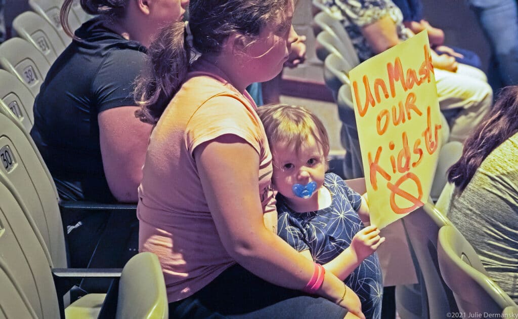 A toddler with a pacifier holds a sign reading 'UnMask our kids' and leans against a young woman seated in an auditorium.