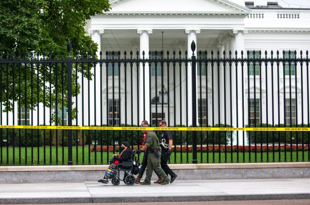 An Indigenous woman in a wheelchair is pushed by two policemen in front of a fence and the White House