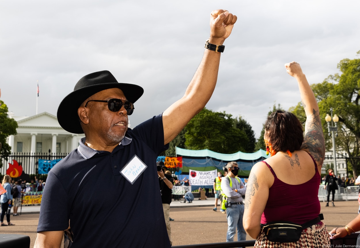 A Black man in a blue t-shirt and black wide-brim hat raises his fist, alongside a white woman in tank top also raising her fist, outside the White House.