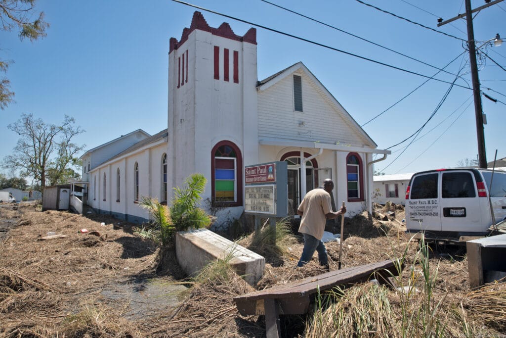 A man walks in front of a damaged white church with a bell tower on a sunny day