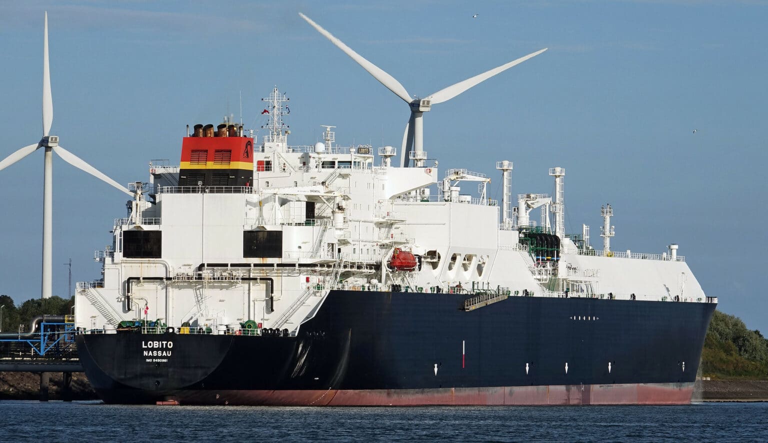 Large LNG tanker vessel passing in front of two white wind turbines