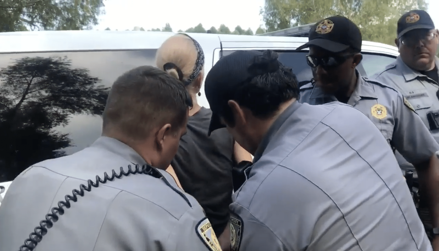 A blonde woman is pressed against an SUV while three men in gray law enforcement uniforms handcuff her, with a fourth looking on
