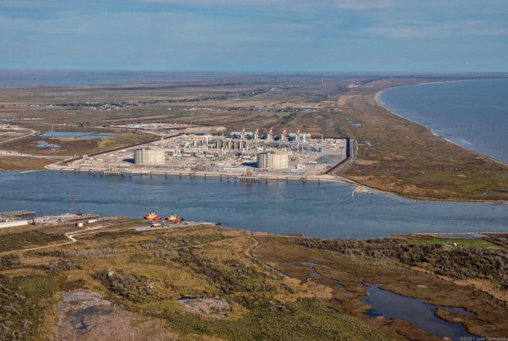 Aerial view of a large industrial facility with piping and tanks on flat marshy land and water on two sides