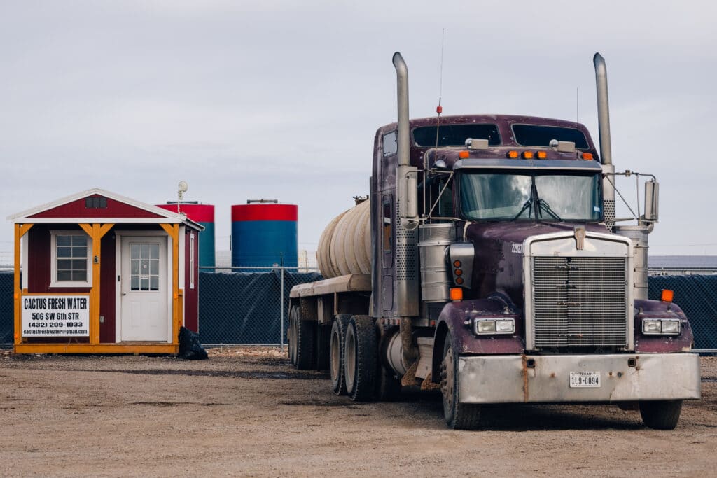 Purple semi truck with a white tank parked on dirtpan next to two red-capped blue tanks and a kiosk with the sign 'Cactus Fresh Water Sales'