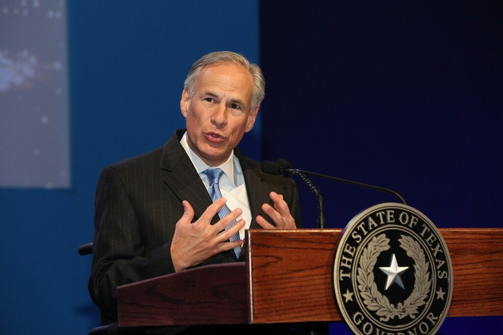 Man with gray hair speaking with his hands partially raised while at a wooden podium with The State of Texas Governor seal on front