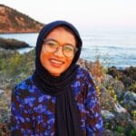 Young woman with glasses smiling, wearing a black hijab and black and purple shirt, with ocean and hills behind her