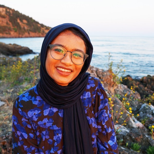 Young woman with glasses smiling, wearing a black hijab and black and purple shirt, with ocean and hills behind her