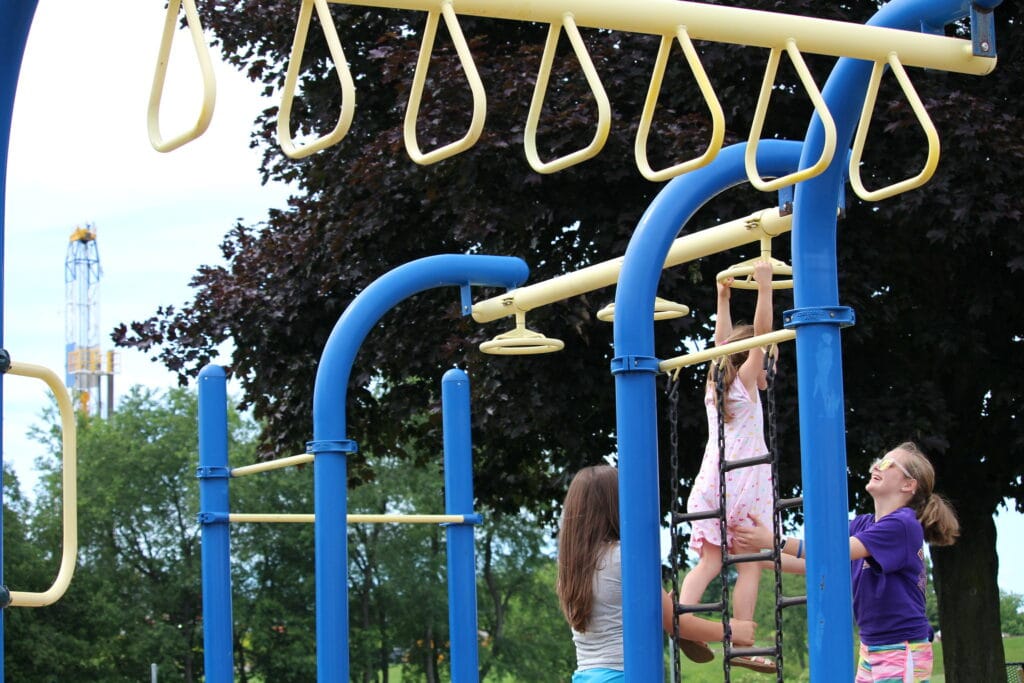 Women helping child at monkey bars with drilling tower visible behind trees