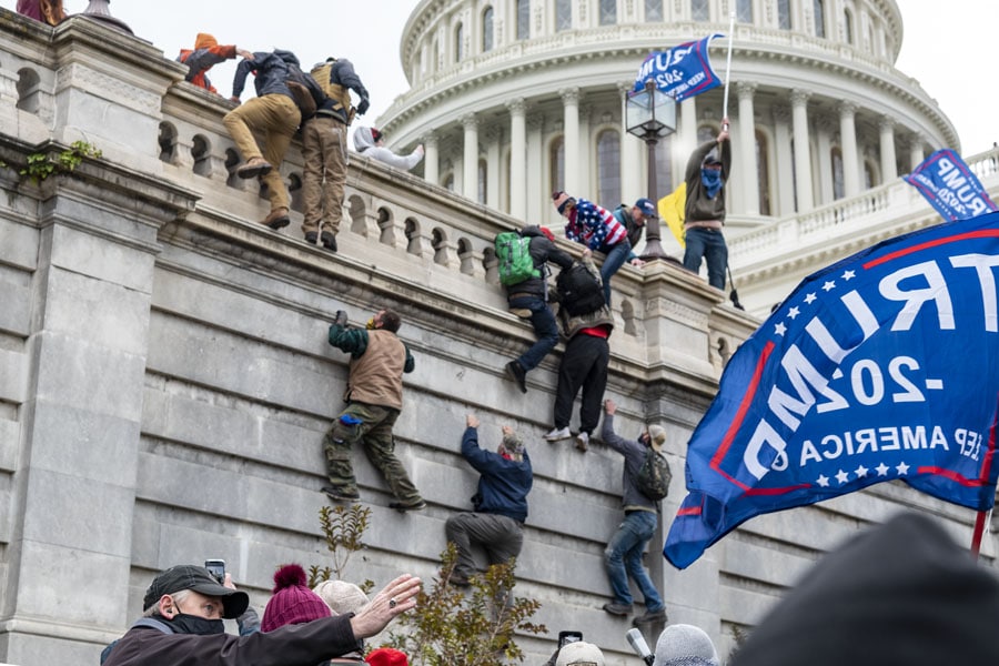 Men scale the stone facade of the U.S. Capitol while others wave large blue Trump 2020 flags