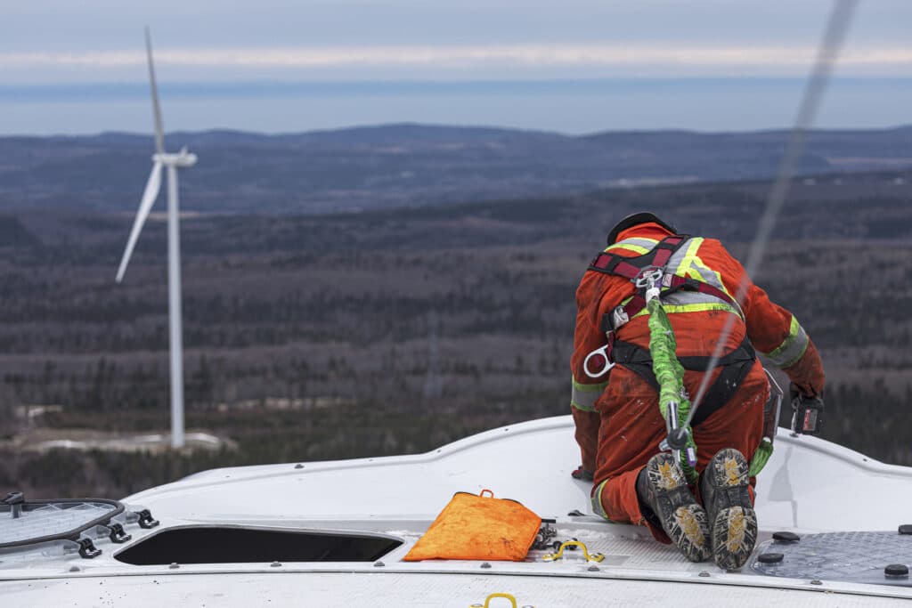 Worker in orange jumpsuit and safety harness tightens bolts on top of a white wind turbine, with another turbine visible in the distance