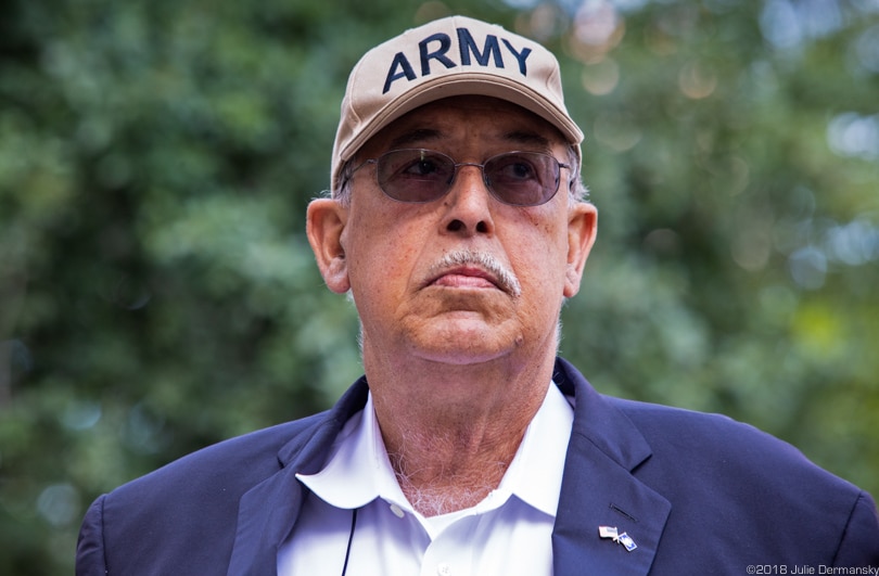 Headshot of an older man with white mustache, blue blazer, tinted glasses, and tan baseball cap reading 'ARMY'