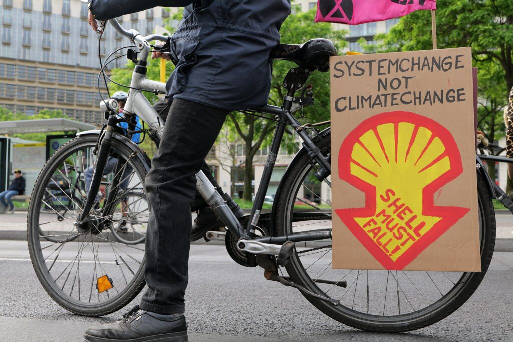 Lower half of person in black standing on their black bicycle in downtown Berlin with a cardboard sign painted with 'systems change not climate change' and an adapted yellow and red Shell logo pointing down with 'Shell must fall', hanging on the back wheel