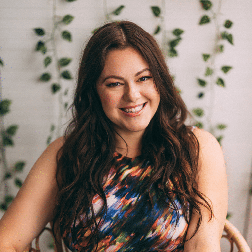 Woman with long dark brown hair, wearing a multi-colored dress, smiles at the camera, with green vines on a white wall behind her