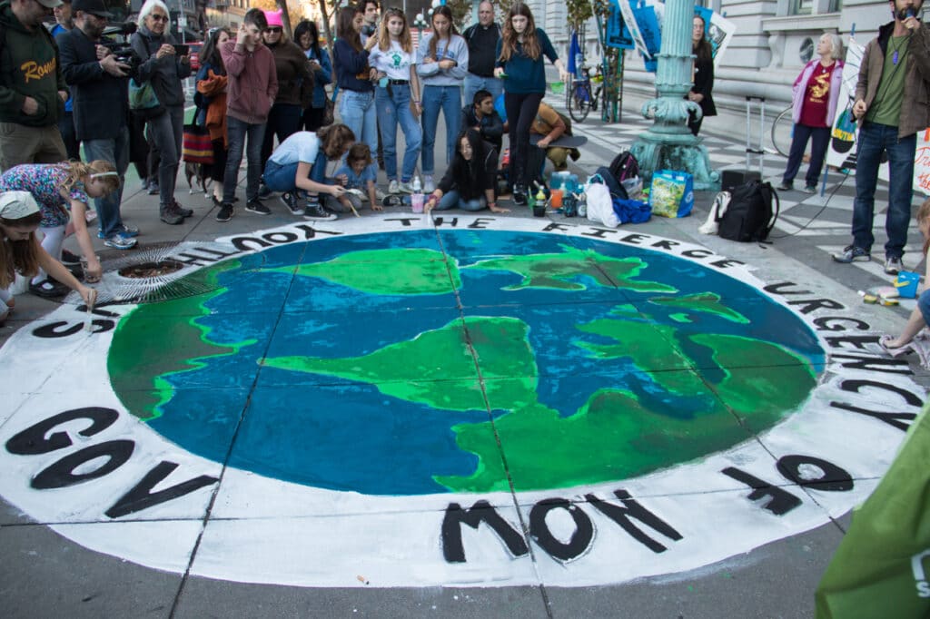 Children painting a mural of the Earth, surrounded by a white band with black text 'Fierce urgency of now, Youth v Gov,' on the sidewalk outside a courthouse with adults watching
