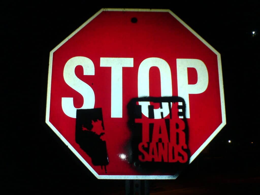A red and white stop sign with a black painted stencil on its bottom half, so that it now reads "Stop the Tar Sands."