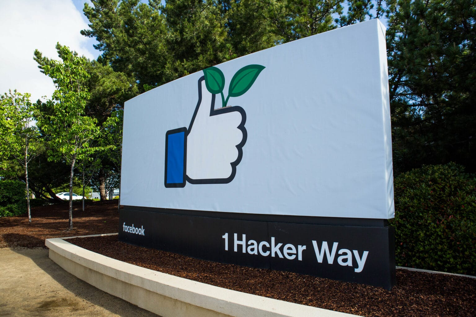 A sign at Facebook's headquarters shows the site's "thumbs up" image holding a leafy green plant, and the words 1 Hacker Way.