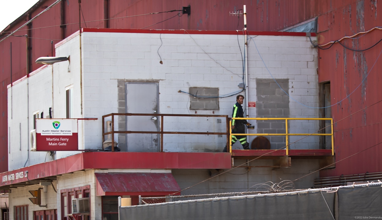 A man walking on a second story balcony outside a large white and red building. A sign reads 'Austin Master Services - Martins Ferry Main Gate'