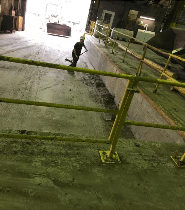 A person with bare forearms and wearing a hardhat holds a push broom in a dirty cement-floor warehouse, next to a fenced ramp. A backhoe is visible just outside an open garage door.