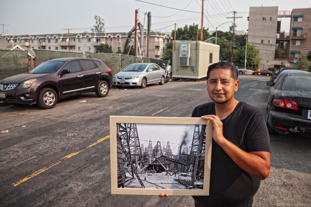Man in black shirt holds framed black-and-white photo of oil derricks, a road with cars, apartments, and construction in background.