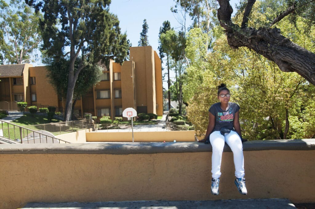 A middle school-aged Black girl sits on a wall by a basketball court and apartment