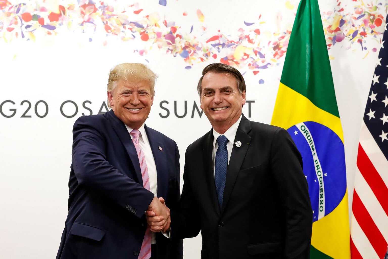 Donald Trump shakes hands with Jair Bolsonaro with a Brazilian flag