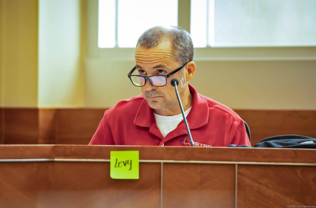David Levy, dressed in a red polo shirt, looks down his glasses  from behind a microphone and wood paneled dais with a line green sticky note that reads 'Levy.'