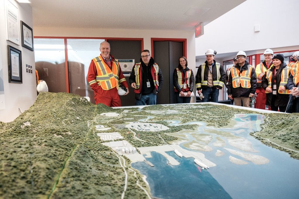 Premier John Horgan in orange safety vest standing next to LNG Canada 3D project map with men and women in safety vests and hard hats