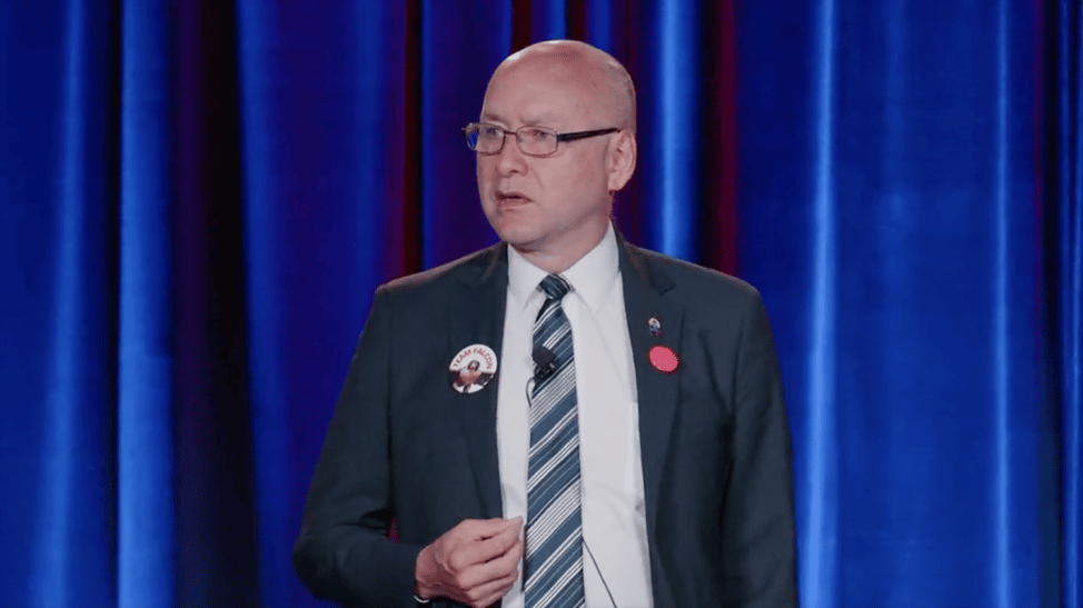 Bald man in glasses and suit shown from waist up standing in front of a blue stage curtain