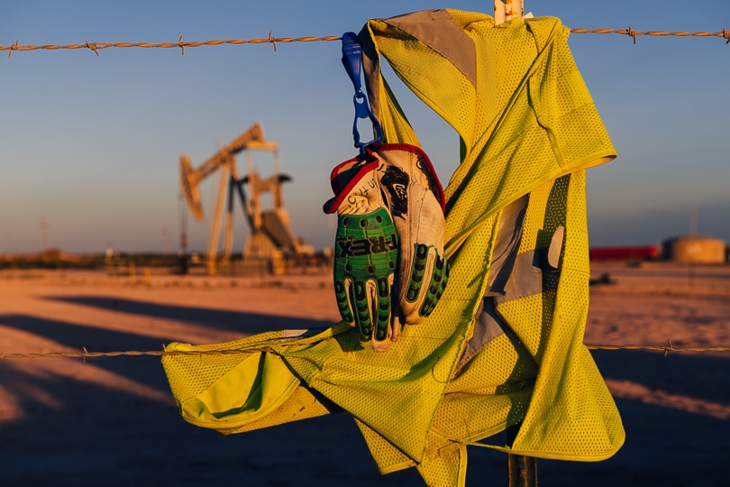 A yellow oilfield worker's vest and gloves hang on a fence post in front of an idled pump jack