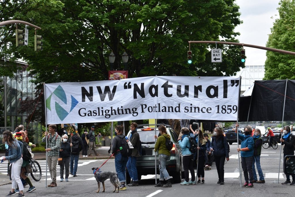 Youth protesters cross a street carring a white banner reading 'NW "Natural" Gaslighting Portland since 1859"'