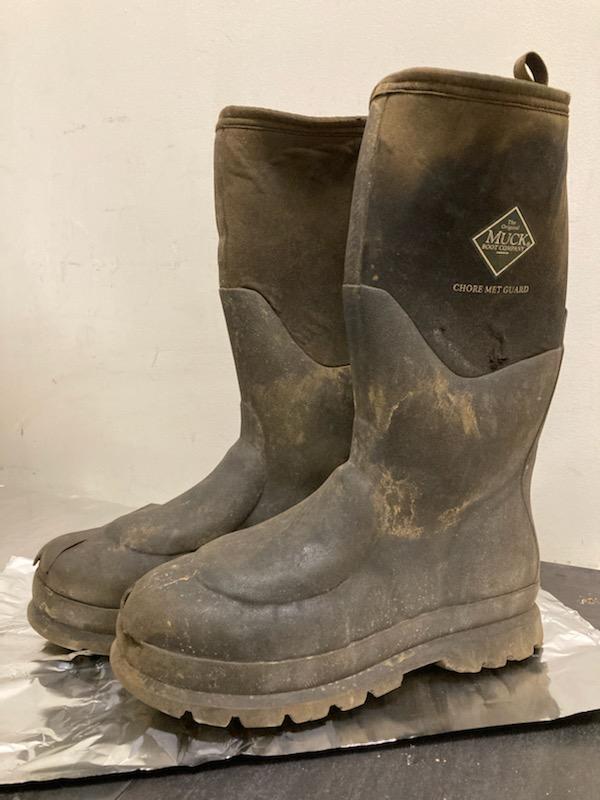 Black pair of muck boots with visible debris dusted over most of the boots, which are sitting on aluminum foil against a white background