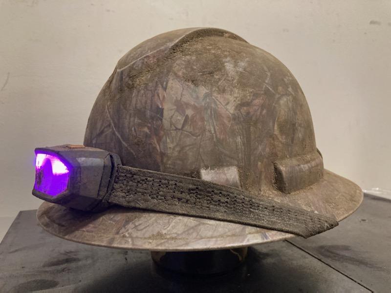 A dirty-gray hard hat with short brim and headlamp with purple light, sitting against a white background.