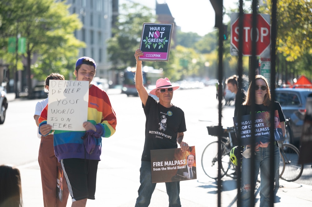 Four young activists hold signs calling David Malpass a climate denier and calling for his resignation, on a DC city street outside the World Bank headquarters